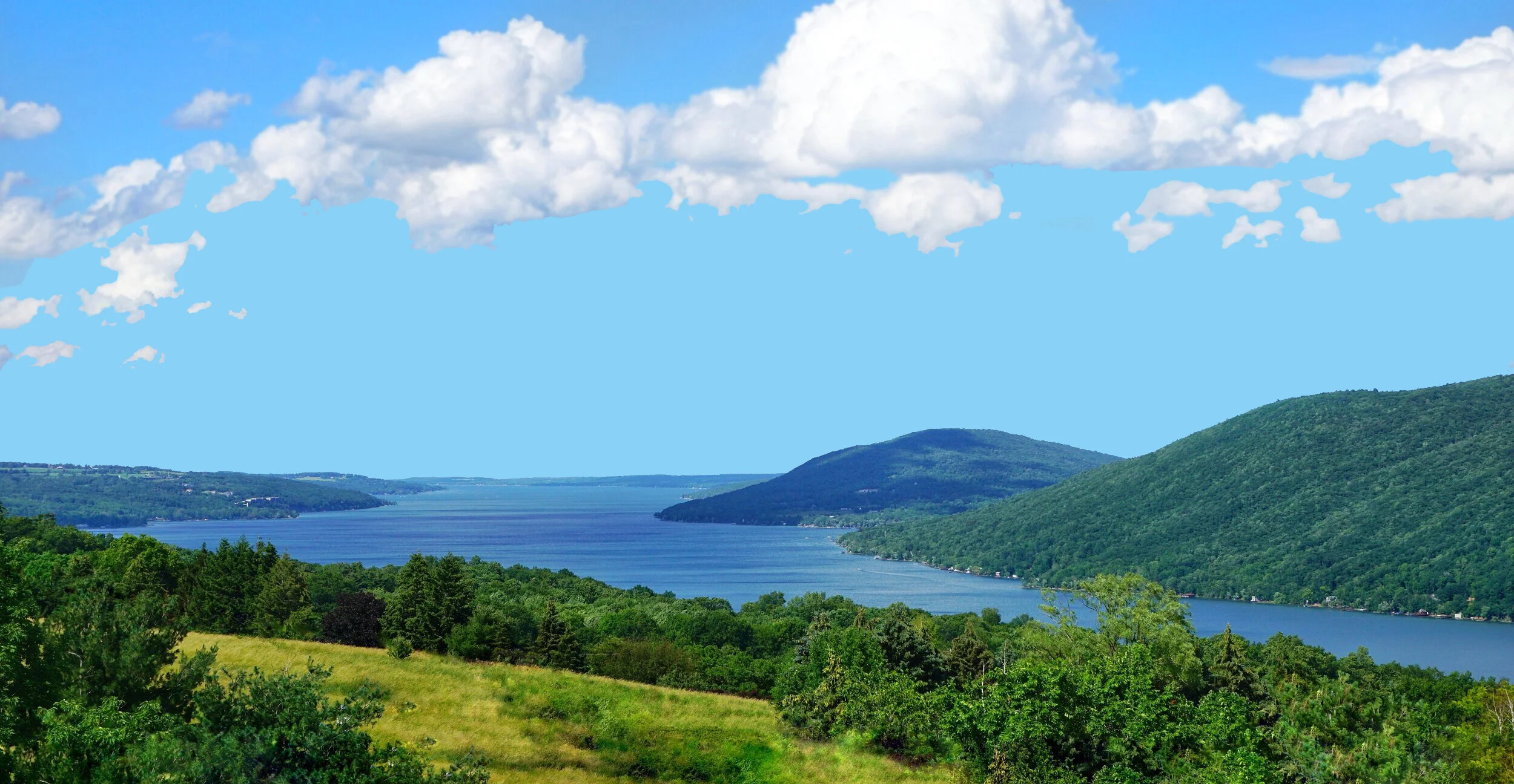 A scenic view of Canandaigua Lake, part of the Canandaigua Lake Wine Trail, surrounded by green hills and forests under a bright blue sky with fluffy white clouds.