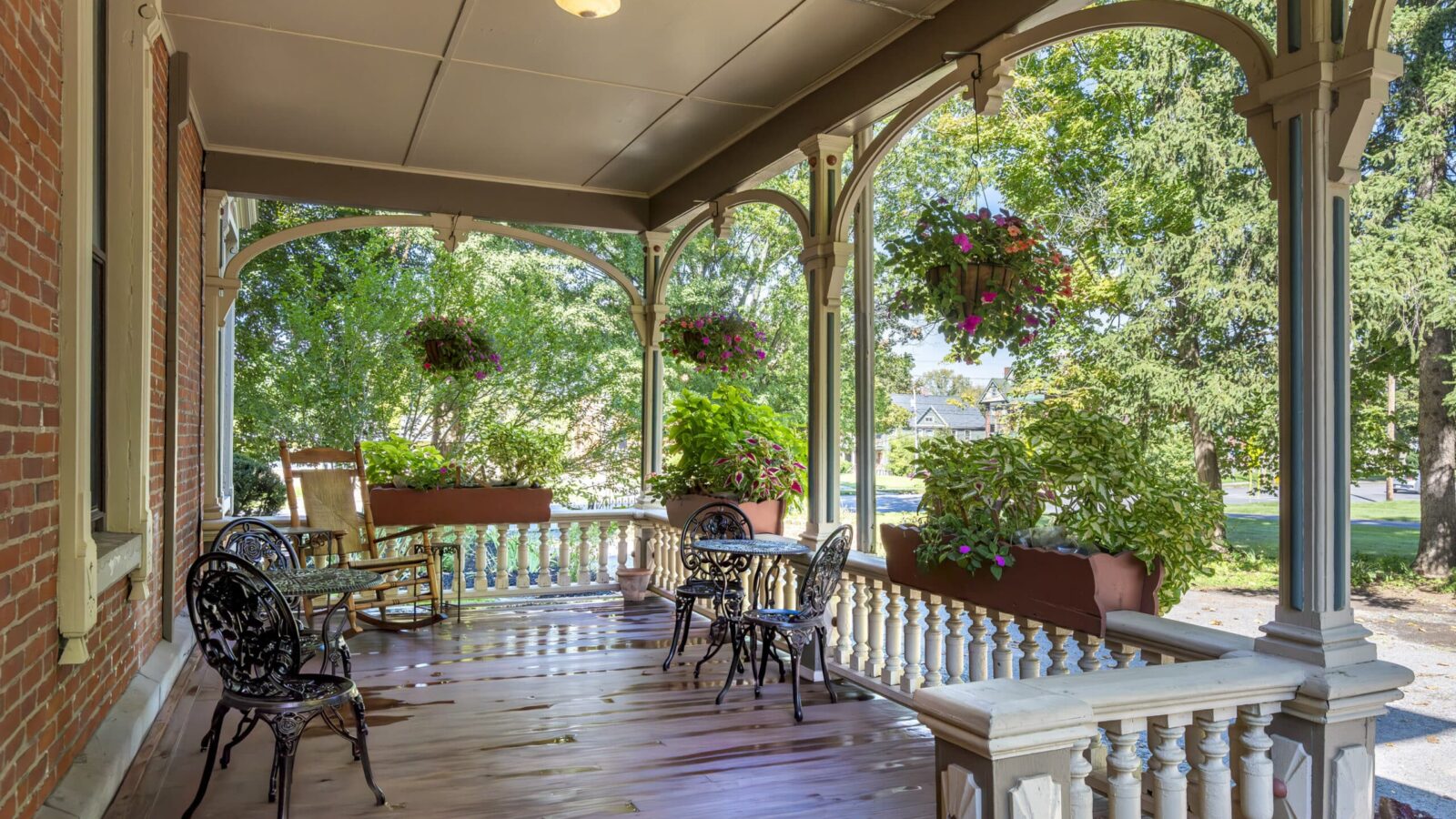 A wooden porch at Rosemont Inn Bed & Breakfast features black metal chairs, a table, a rocking chair, hanging flower baskets, and potted plants, all overlooking a green yard with trees on a sunny day.