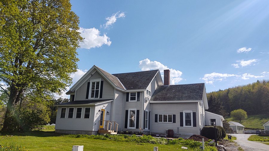 White two-story farmhouse with dark roof and a small porch, surrounded by green grass and trees under a bright blue sky with scattered clouds—a welcoming setting reminiscent of The Paw House Inn.