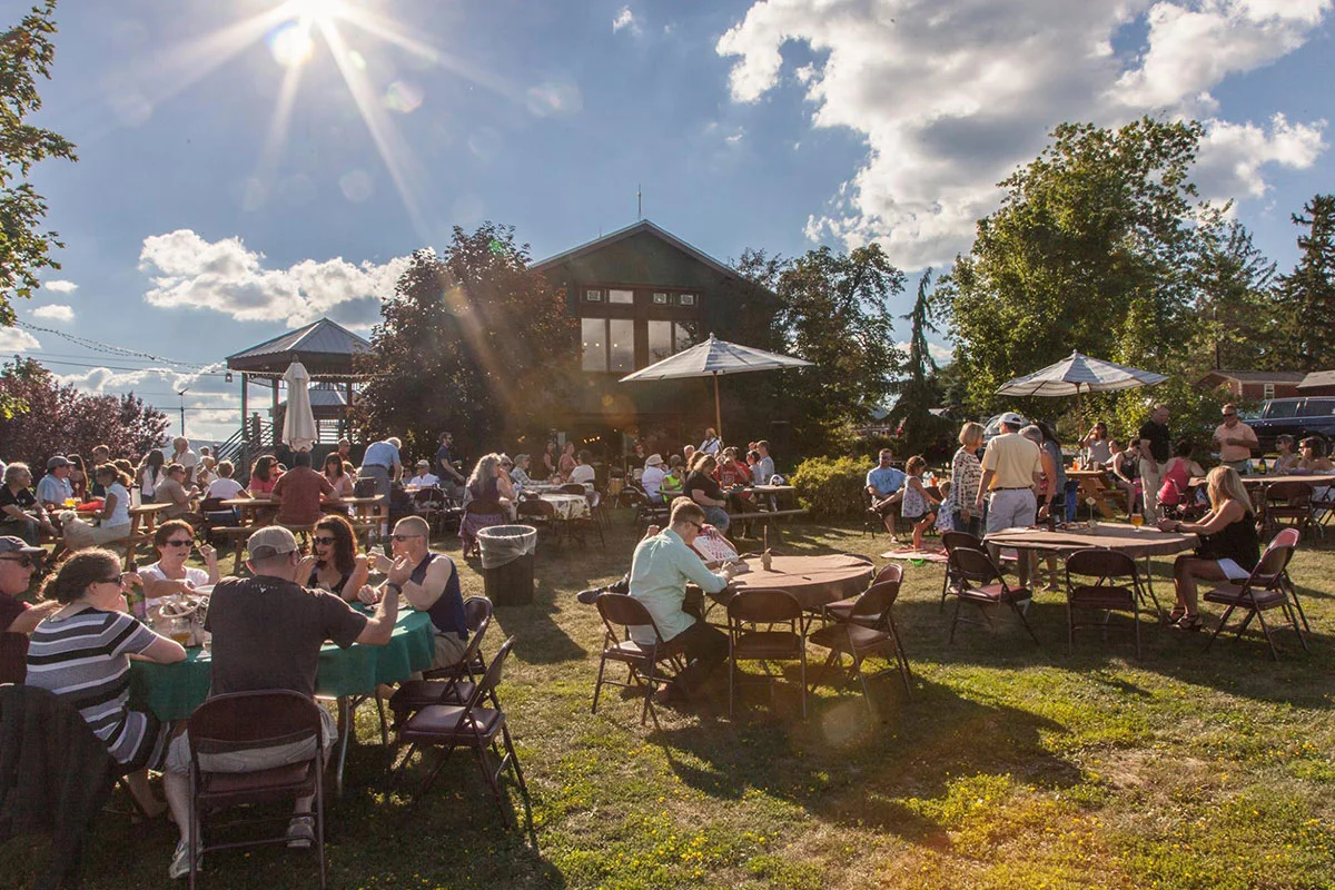 People gather at outdoor tables on a sunny day, enjoying food and conversation along the Canandaigua Lake Wine Trail. Umbrellas provide shade, and a house or barn is visible in the background among trees and clouds.