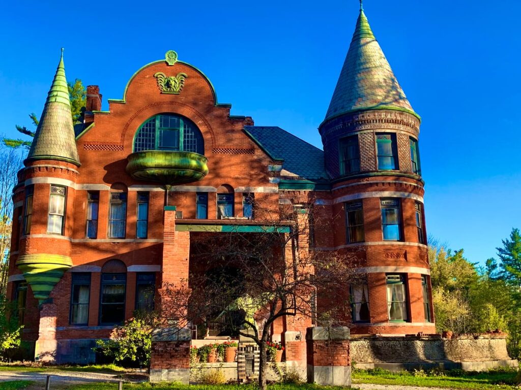 A large, historic red brick mansion reminiscent of Wilson Castle, with two round towers, conical roofs, and ornate green architectural details, set against a clear blue sky and surrounded by trees and shrubbery.
