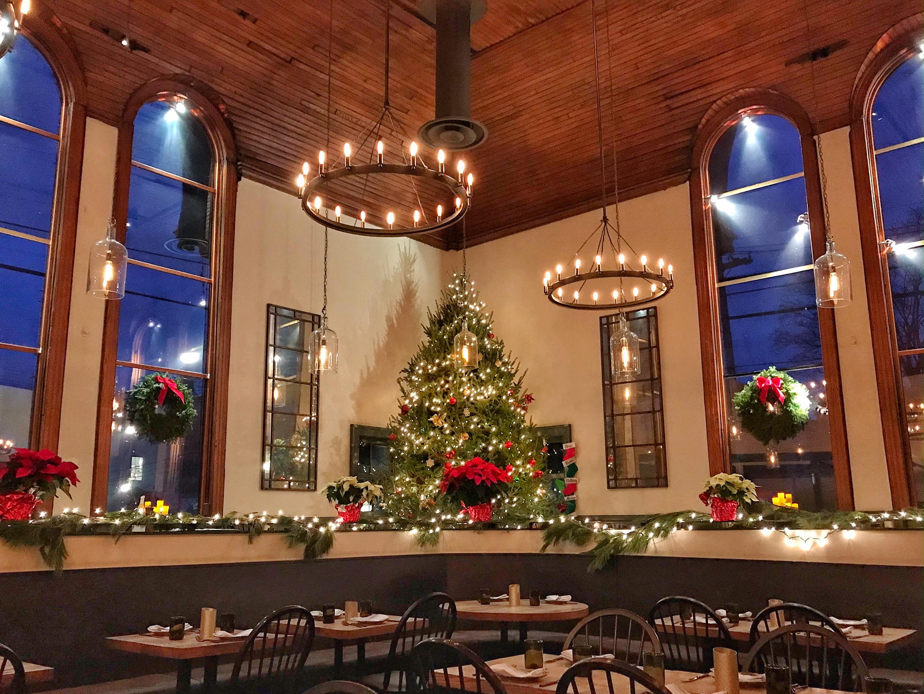 A warmly lit Terrapin Restaurant decorated for Christmas with a large tree, wreaths, poinsettias, and string lights. High arched windows and candle chandeliers create a festive, cozy atmosphere. Tables are set for dining.