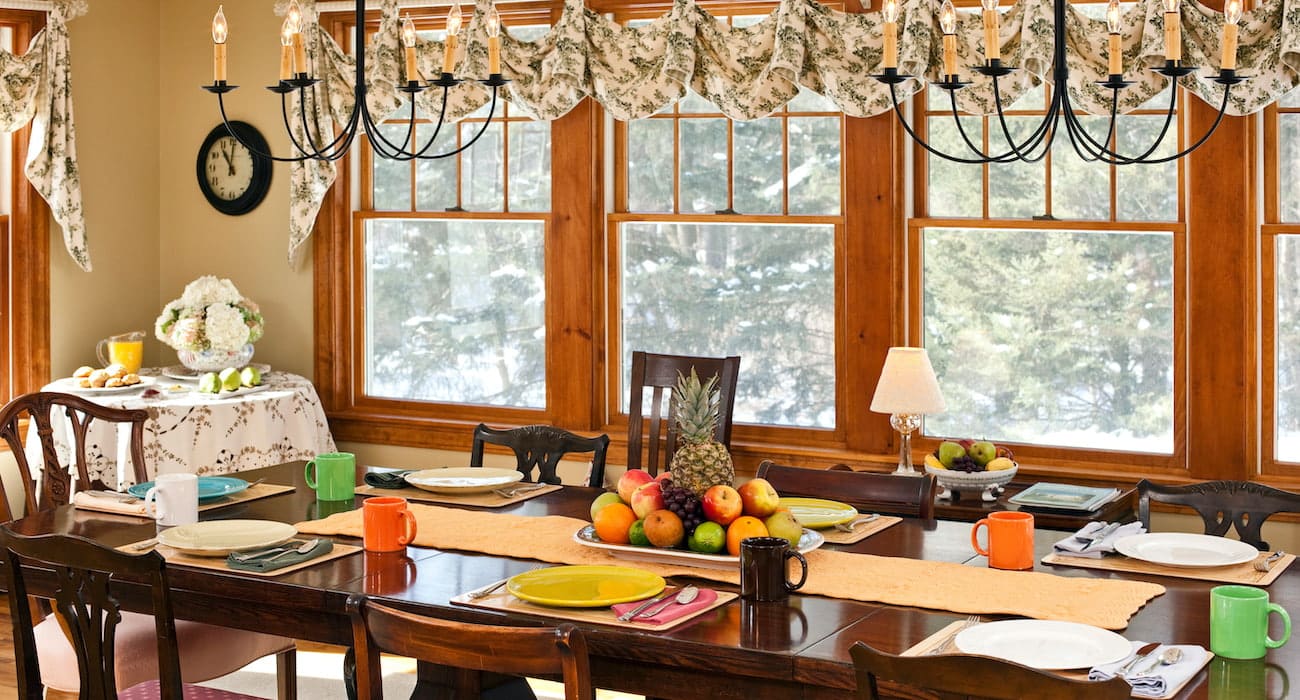A dining room at Journey Inn Bed & Breakfast features a long table set for eight, colorful mugs and plates, a charming centerpiece of fruit, large windows with valances, and two black chandeliers hanging above. Snowy trees are visible outside the windows, creating a cozy and inviting ambiance.