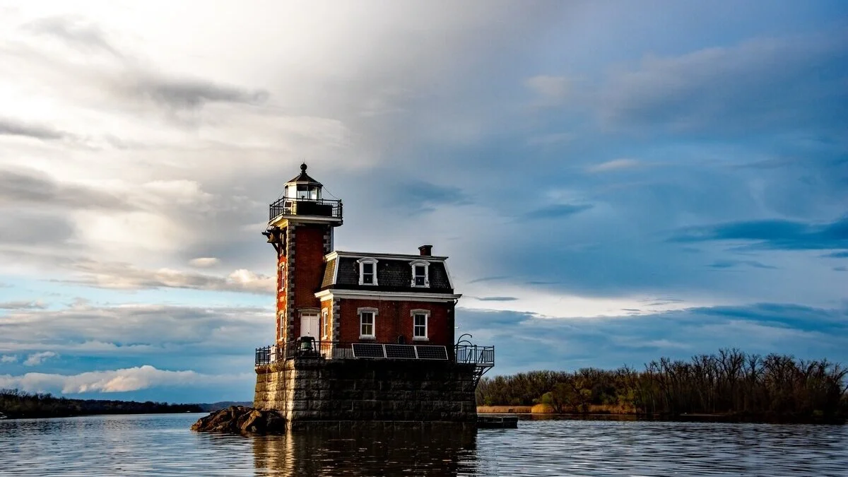 A small brick lighthouse stands on a stone base in the middle of a calm body of water, with trees and a cloudy sky in the background—a tranquil scene reminiscent of the peaceful ambiance found at Journey Inn Bed & Breakfast.