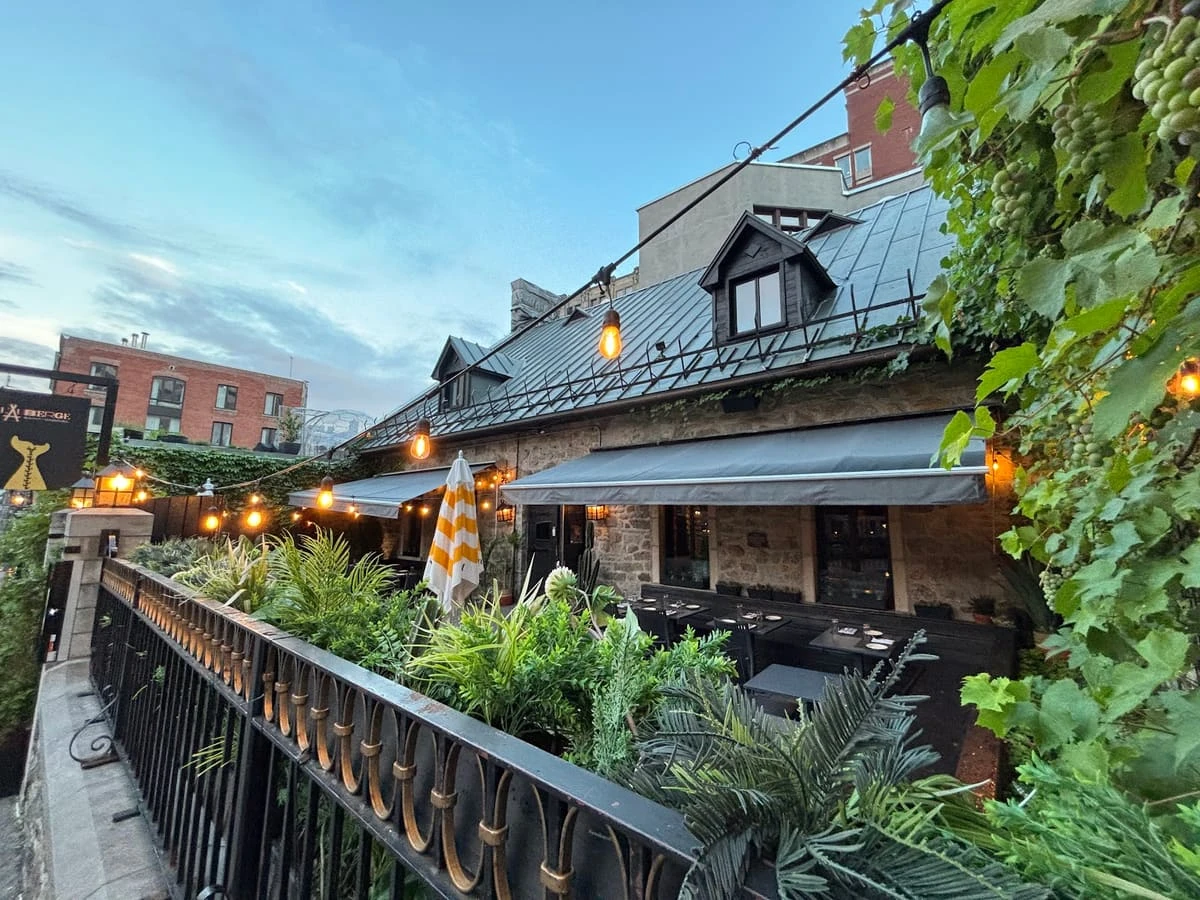 A cozy outdoor restaurant patio with greenery, string lights, black tables, a striped umbrella, and a stone building with dormer windows at dusk—reminiscent of the charming ambiance found at Auberge Saintlo Montréal.