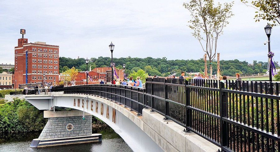 People walk across a curved bridge labeled Amsterdam over a river in Riverlink Park, with trees, lamp posts, and brick buildings in the background on a cloudy day.