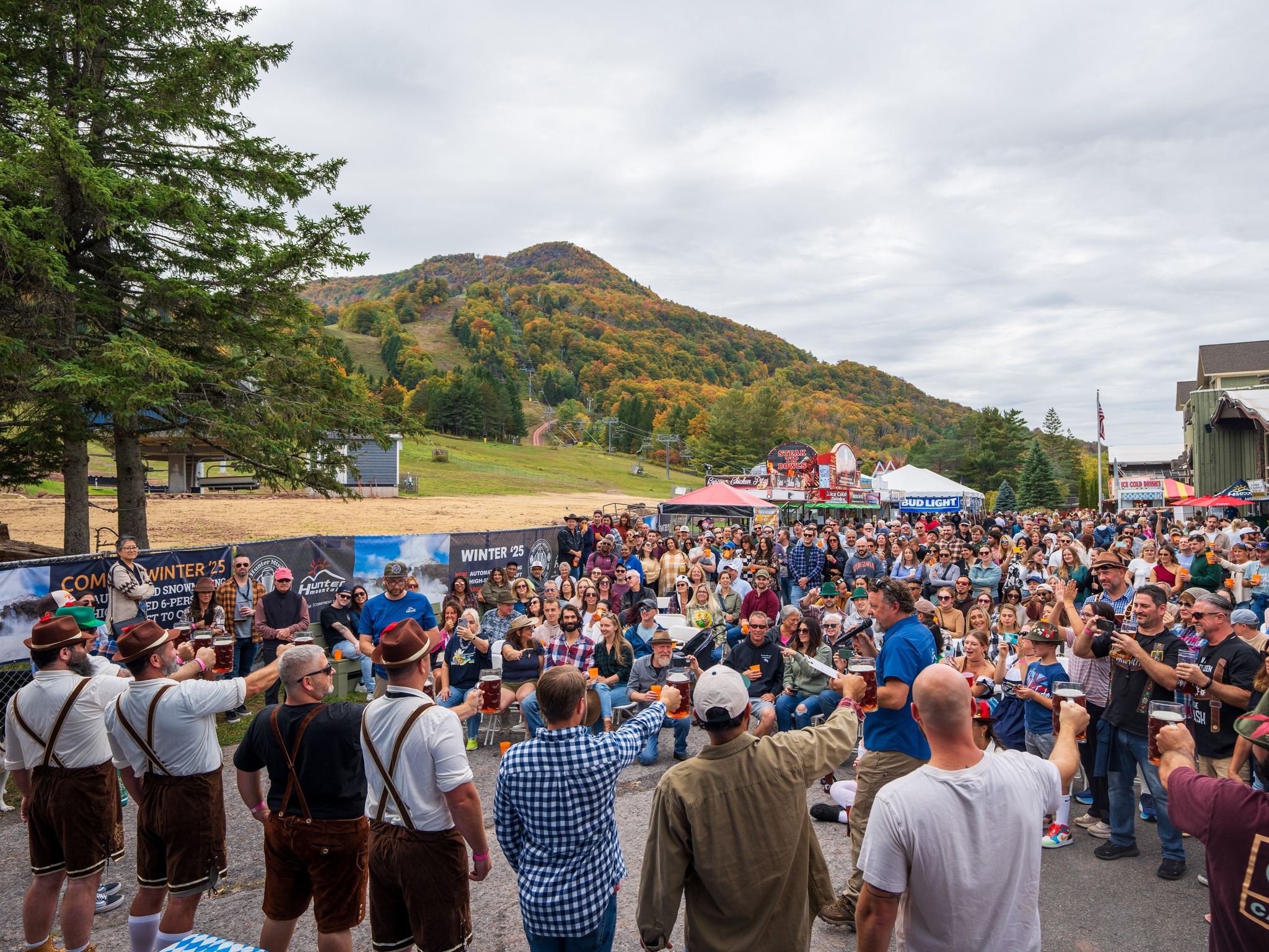 At the Saratoga Fall Festival, a large crowd gathers outdoors at a mountain celebration. People hold up drinks, some wear traditional Bavarian outfits, and autumn trees color the hillside in the background. Booths and tents line the event area.