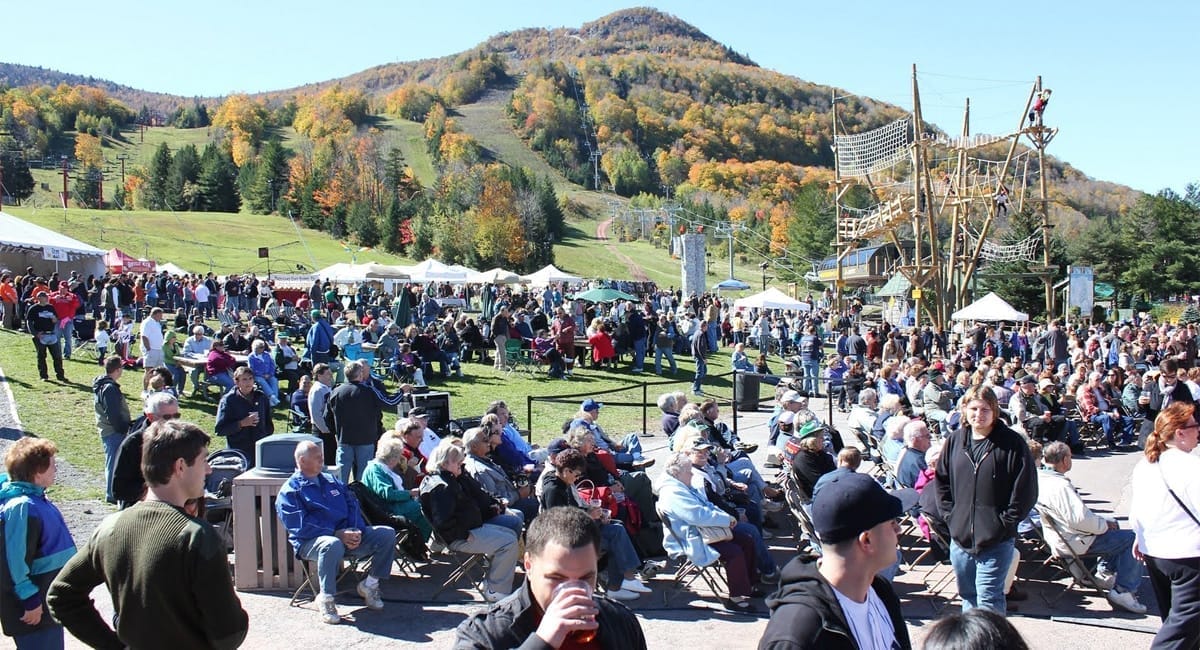 A large crowd gathers outdoors by tents and a climbing structure, with colorful autumn trees and a hill in the background. Many people are seated or walking, enjoying the lively atmosphere of Oktoberfest at Hunter Mountain under the sunny sky.