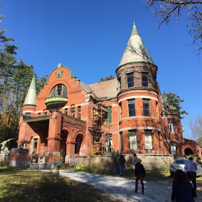 A large red-brick Victorian mansion with turrets and arched windows, reminiscent of The Otesaga Resort Hotel, stands surrounded by trees. Several people walk toward the entrance, and scaffolding is visible on one side of the building.