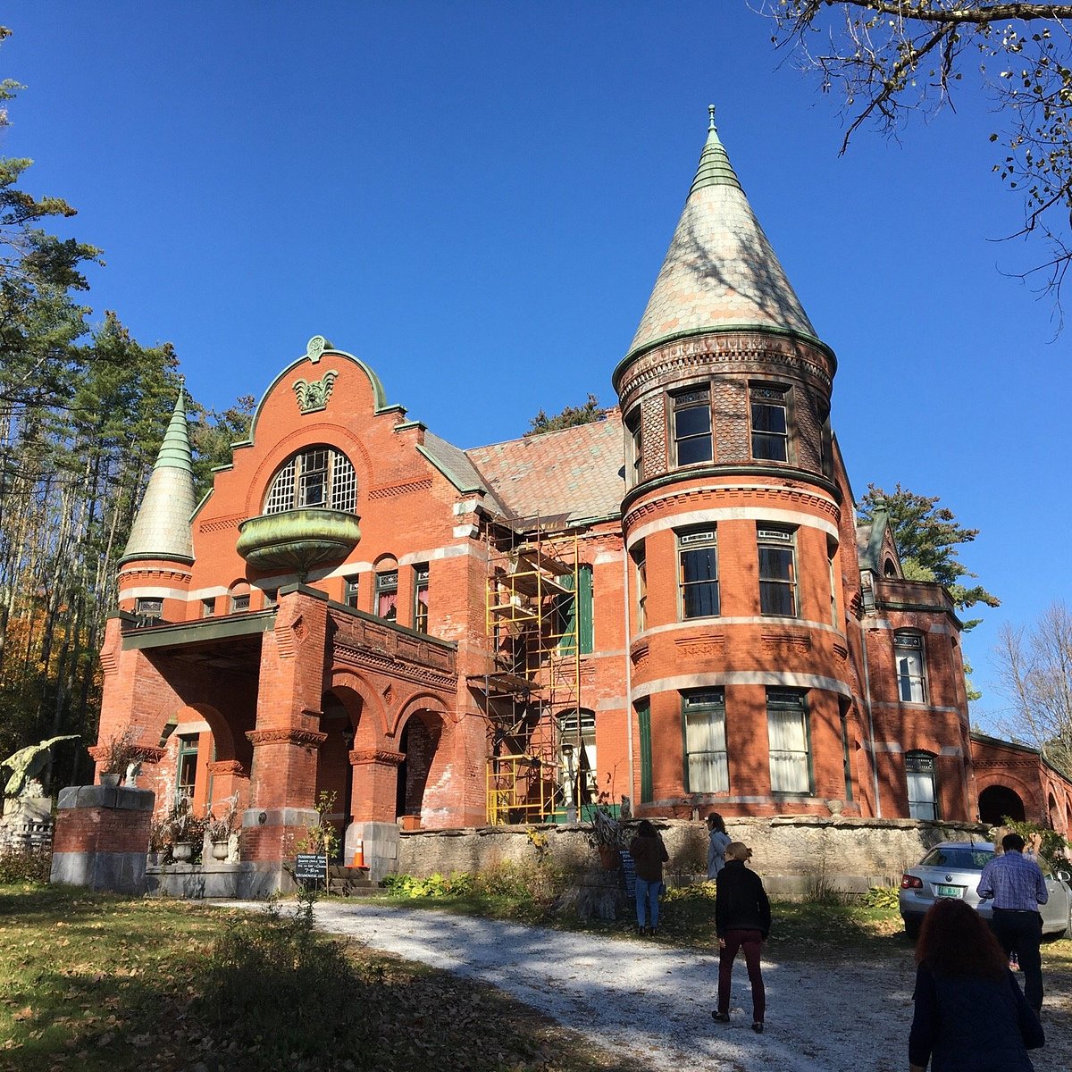 A large red-brick Victorian mansion with turrets and arched windows, reminiscent of The Otesaga Resort Hotel, stands surrounded by trees. Several people walk toward the entrance, and scaffolding is visible on one side of the building.