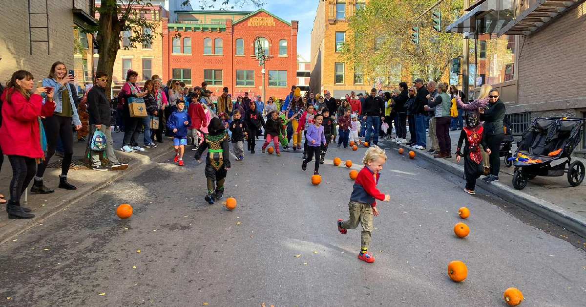 Children run down a city street scattered with pumpkins while adults watch and take photos, capturing the lively spirit of the day. People are dressed in colorful clothes and costumes, suggesting a festive fall or Halloween event that coincides with the city’s annual Wine and Food Festival.