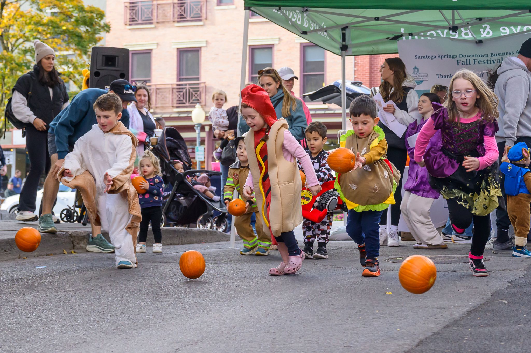 Children in costumes race small pumpkins on a street during the Saratoga Fall Festival, with adults and strollers in the background under a green tent.