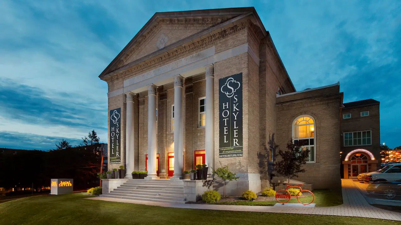 Hotel Skyler, reminiscent of the inviting charm found at Auberge Saintlo Montréal, is a historic brick building with tall white columns, large banners, and warmly lit windows. Pictured at dusk with a red bicycle near the entrance and a lit sign on the lawn, it offers both character and comfort in every detail.