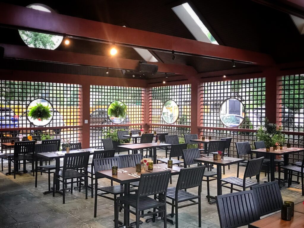 Spacious patio dining area with black tables and chairs, circular windows, potted plants, and skylights letting in natural light—perfect for relaxing and enjoying the serene atmosphere near Riverlink Park. No people are present.