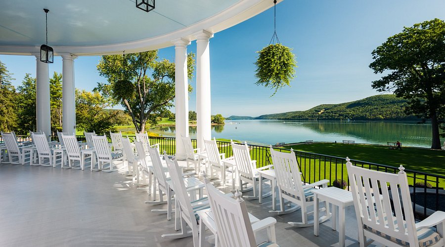 A porch with white rocking chairs overlooks a calm lake, lush green lawn, and distant hills under a clear blue sky. Columns and a hanging plant add charm to the peaceful, scenic setting—reminiscent of the timeless elegance found at The Otesaga Resort Hotel.