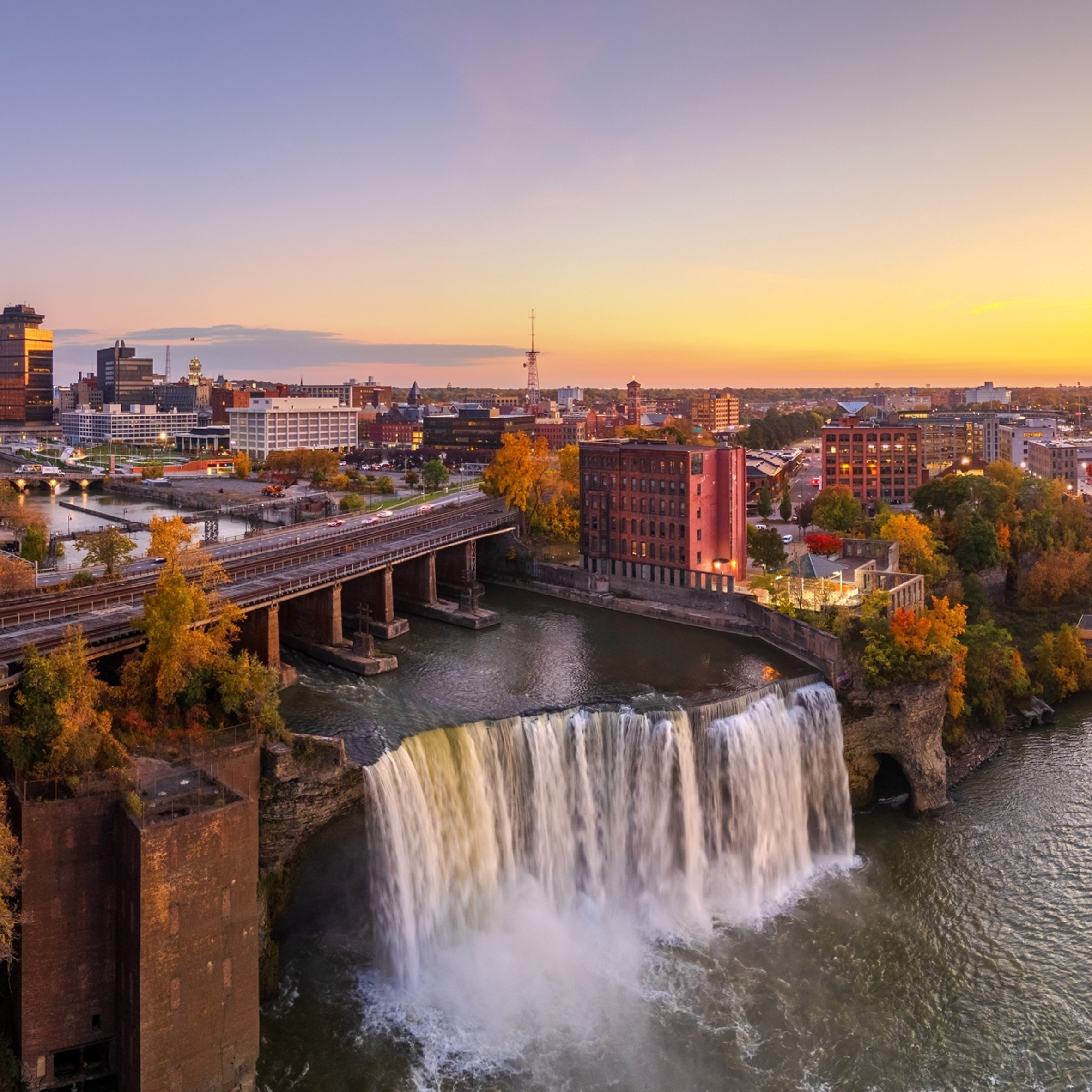 High Falls in Rochester at sunset, with city buildings and a bridge in the background, a scenic stop along the Adirondack Craft Beverage Trail.